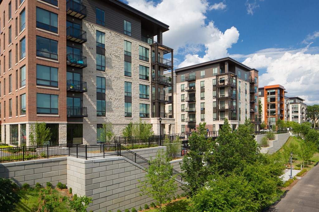 Modern Inspire Apartment Homes buildings with brick and glass facades are surrounded by lush greenery under a bright blue sky with scattered clouds.