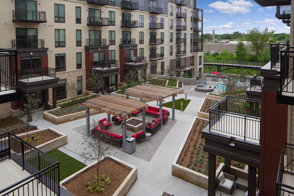 Modern courtyard here at Inspire Apartment Homes with a symmetrical design featuring wooden pergolas, red sofas around fire pits, lush landscaping, and a distant pool area.