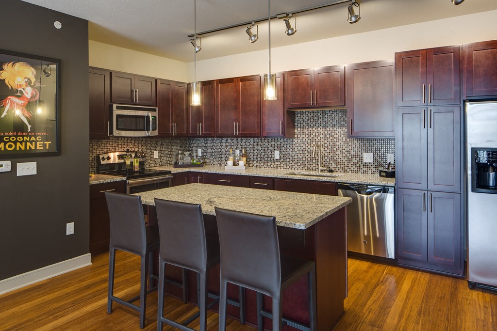 Modern kitchen here at Inspire Apartment Homes with dark wood cabinets, granite countertops, and mosaic backsplash. An island with three brown chairs and stainless steel appliances. Hanging lights create a cozy ambiance.
