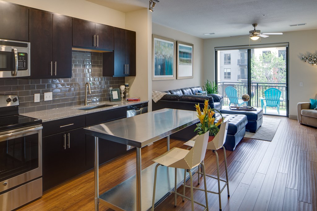 Modern apartment interior here at Inspire Apartment Homes with an open-plan kitchen and living room. Dark cabinets, stainless appliances, large plant, leather sofa, and balcony with blue chairs.