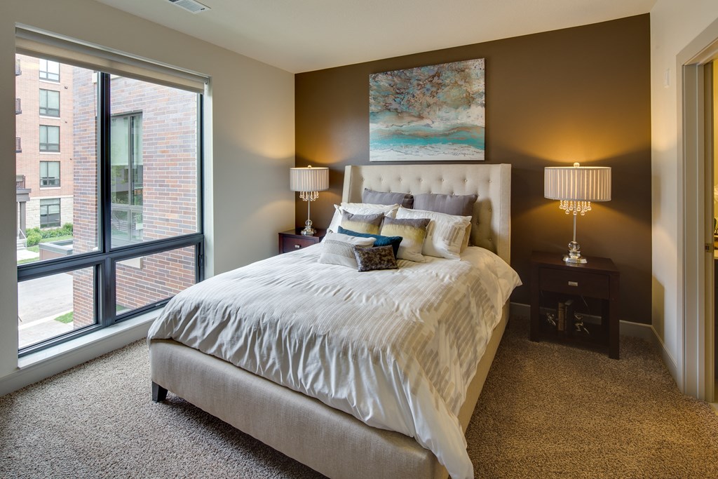 Elegant bedroom here at Inspire Apartment Homes with a plush bed and assorted pillows, flanked by bedside tables and lamps. Large window shows a cityscape, creating a cozy ambiance.
