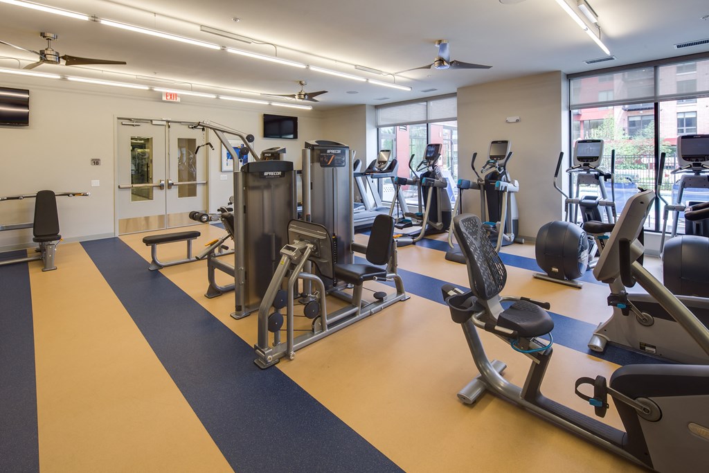 Bright gym interior here at Inspire Apartment Homes with various exercise machines, including ellipticals and weights. Large windows let in natural light, creating an inviting atmosphere.