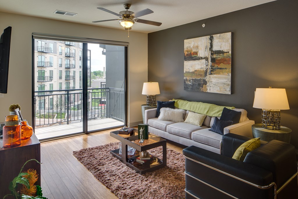 Modern living room here at Inspire Apartment Homes with a gray accent wall, abstract painting, and a white sofa adorned with colorful cushions. Sliding glass doors lead to a balcony with a cityscape view. A ceiling fan and warm lighting create a cozy ambiance.