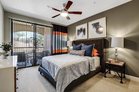 A stylish and inviting bedroom here at Desert Club featuring a spacious bed with layered bedding and accent pillows, flanked by matching nightstands with modern lamps. Large sliding glass doors with vertical blinds and bold drapery panels bring in natural light and provide access to a private patio, while a ceiling fan and soft carpeting add comfort and warmth to the space.