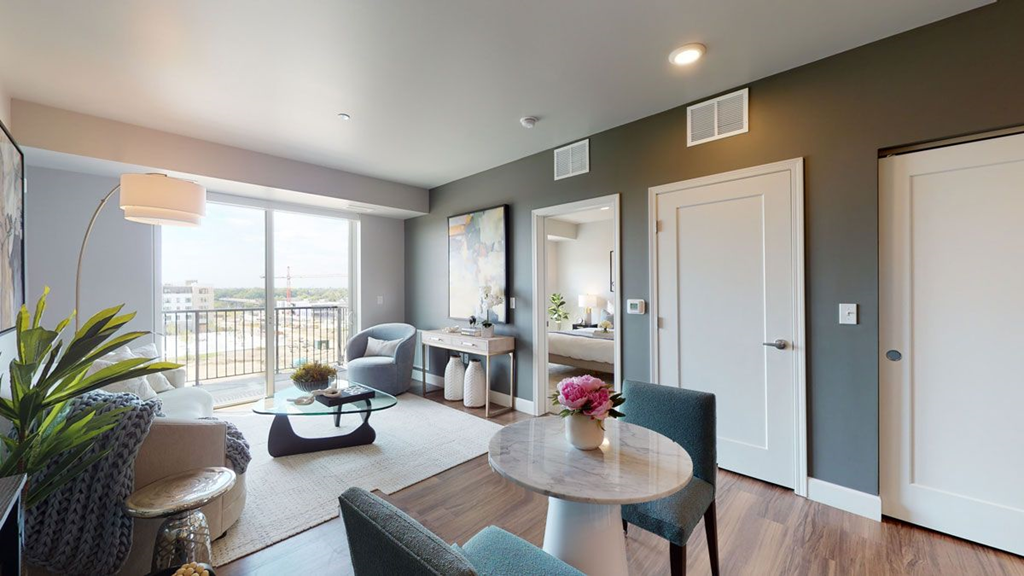 Modern living room here at The Collection at Highland Bridge Apartment Homes with large window, light grey walls, and hardwood floor. Includes a white sofa, green plants, and a round table with floral decor. Bright and inviting atmosphere.