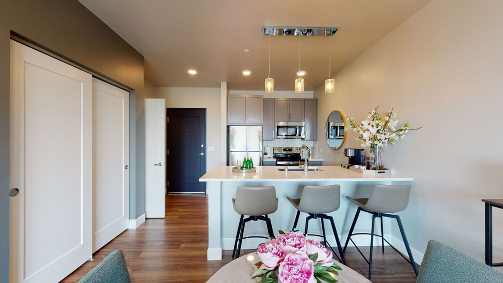 Modern kitchen here at The Collection at Highland Bridge Apartment Homes with sleek gray cabinets, stainless steel appliances, and a beige countertop. Three gray barstools and pink flowers add elegance. Warm lighting creates a cozy ambiance.