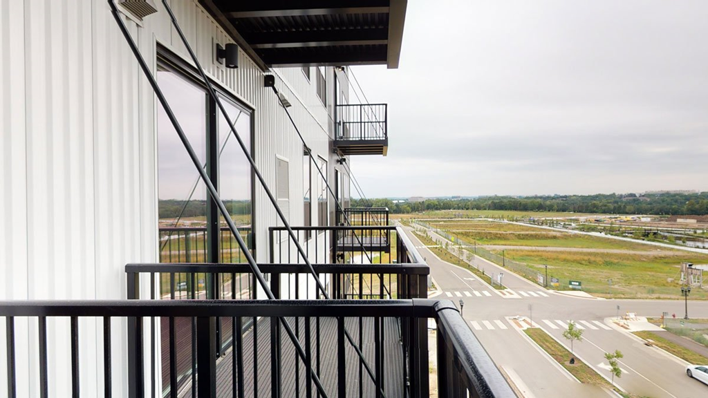 View from a modern apartment balcony here at The Collection at Highland Bridge Apartment Homes showing parallel balconies, a quiet road, grass fields, and distant trees under a cloudy sky, conveying tranquility.