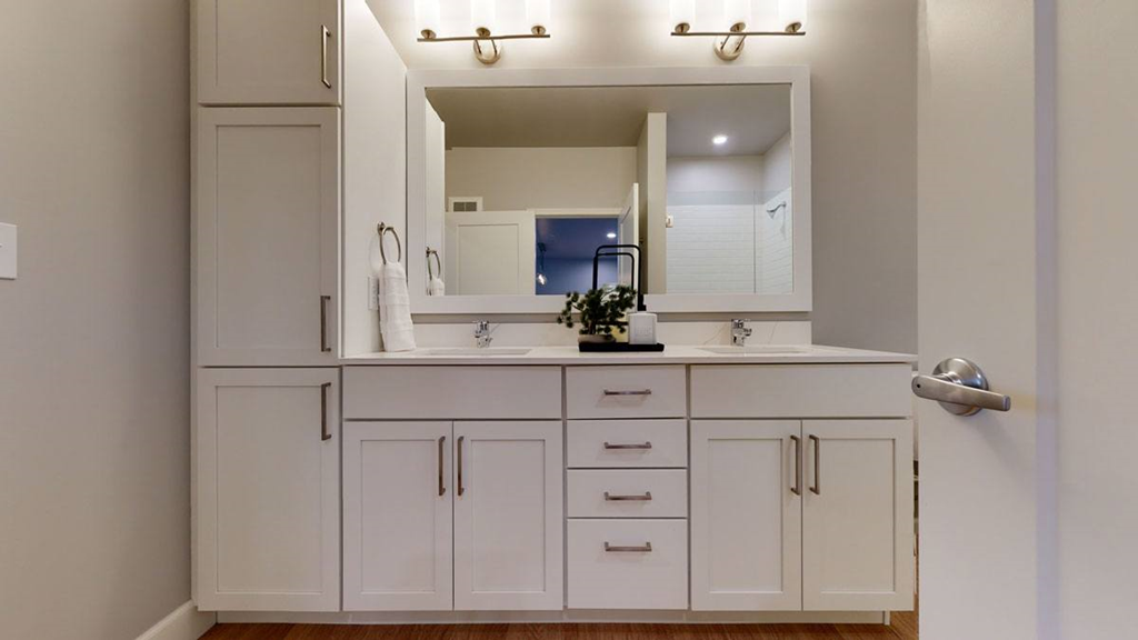 Modern bathroom here at The Collection at Highland Bridge Apartment Homes with dual sinks, large mirror, and white cabinetry. Soft lighting, neat decor, and a towel on a ring holder create a calm ambiance.