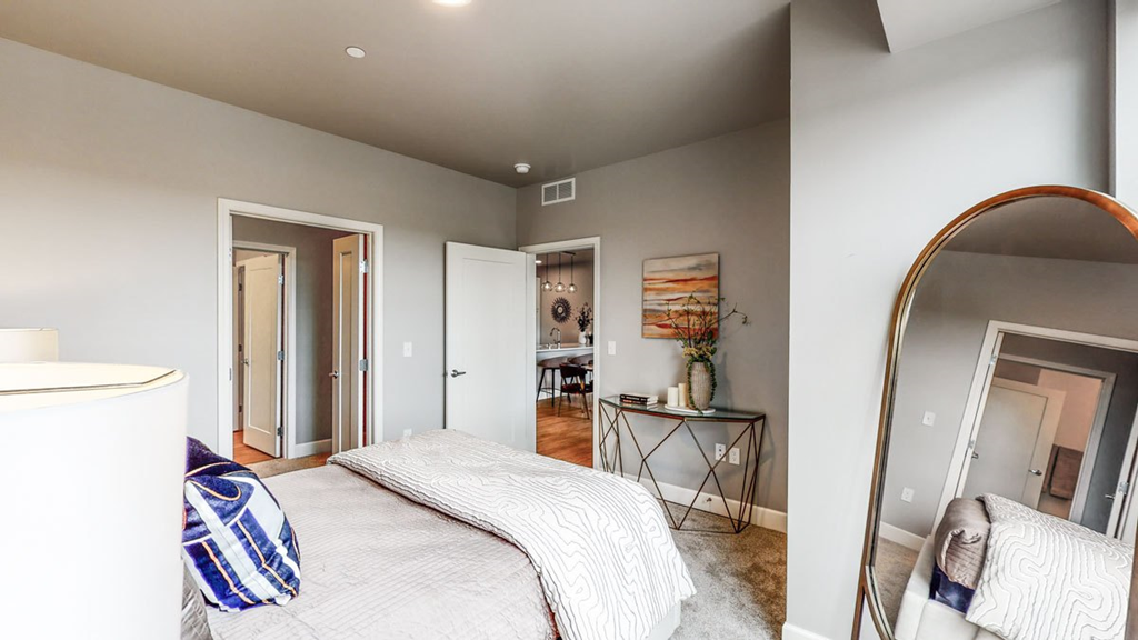Modern bedroom here at The Collection at Highland Bridge Apartment Homes with grey walls and soft lighting. A neatly made bed is in the foreground. A console table with decor and art hangs by a doorway leading to a kitchen.
