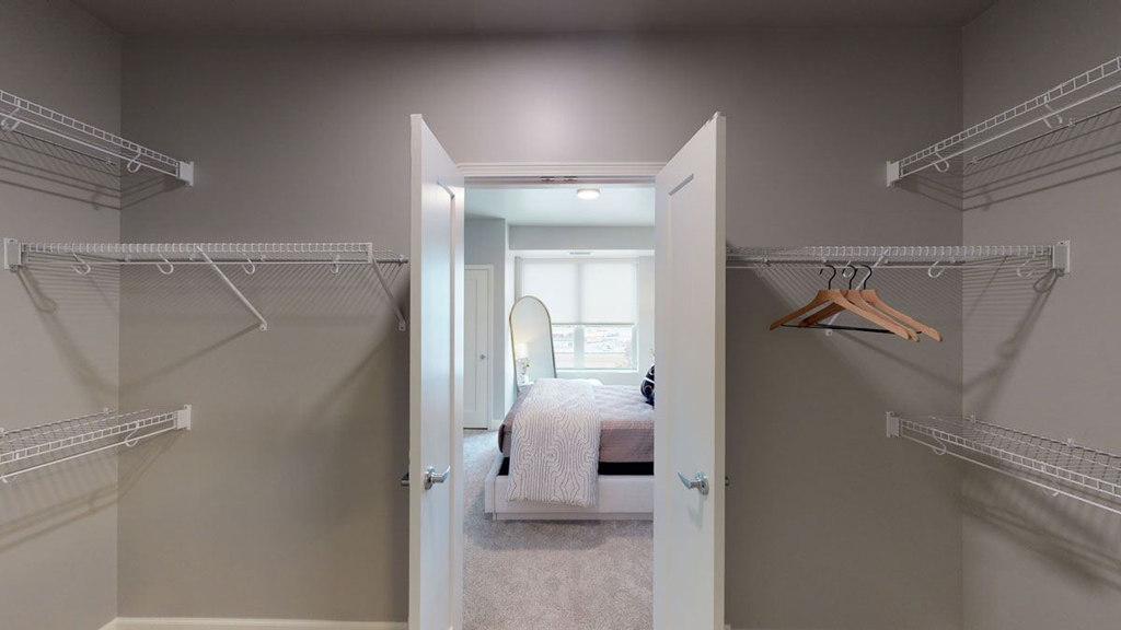 Spacious walk-in closet here at The Collection at Highland Bridge Apartment Homes with white wire shelves and hangers opens to a bright bedroom with a neatly made bed and a large window, creating a tidy atmosphere.