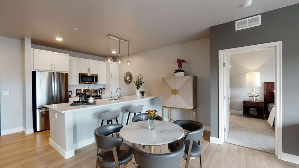 Modern kitchen here at The Collection at Highland Bridge Apartment Homes with a marble island, pendant lights, and gray chairs. Adjacent dining area with a round table. Open doorway to a cozy bedroom.