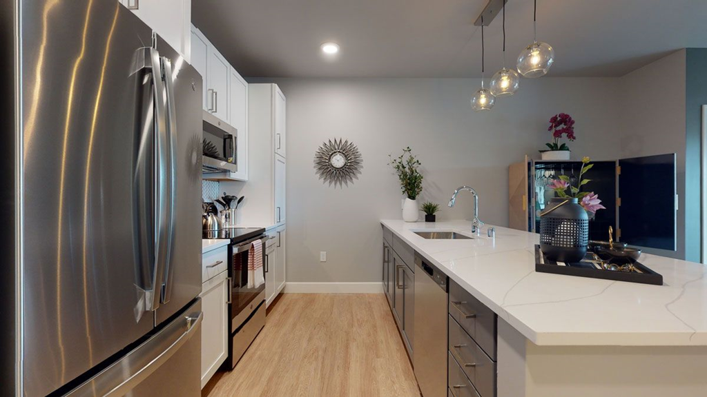 Modern kitchen here at The Collection at Highland Bridge Apartment Homes with stainless steel appliances, white cabinets, a large island with a sink, hanging lights, and decorative plants on the counter.
