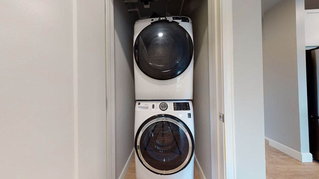Stacked washer and dryer in a narrow laundry closet here at The Collection at Highland Bridge Apartment Homes. The appliances are modern with circular doors, surrounded by light gray walls and light wood flooring.