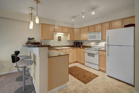 A spacious kitchen here at Admirals Cove with light brown cabinets, granite counter tops, bar type seating, and a ceramic sink. The room has a warm tone.
