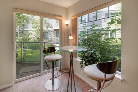 A bright dining nook at Admirals Cove featuring a round glass table with two modern bar-height chairs. Large windows and a sliding glass door overlook green landscaping, filling the space with natural light. Soft carpeting and corner lighting create a relaxed spot for meals or morning coffee.