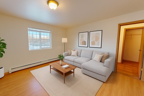 Bright living room at Amber Ridge with wood-style flooring, a large window, and a light gray sectional sofa with neutral pillows. A wooden coffee table sits on a cream area rug, paired with framed abstract art and a floor lamp. An open doorway reveals a walk-in closet, and a potted plant adds greenery.