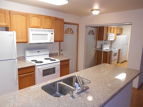 Spacious Amber Ridge apartment kitchen with oak cabinets, white appliances, granite-style countertops, double sink, and breakfast bar overlooking open living area.