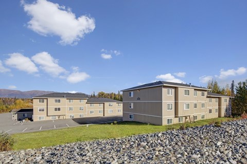 Wide exterior shot of Amber Ridge Apartments showing two beige three-story residential buildings with multiple windows, surrounded by trees with autumn colors and mountains visible in the distance under a bright blue sky.