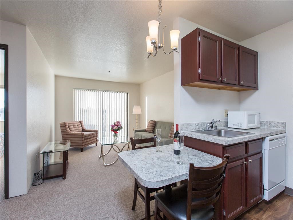 Cozy living area here at Outlook Apartments with striped armchairs, glass table, and flowers. Kitchenette has marble countertops and wine. Bright, relaxed ambiance.