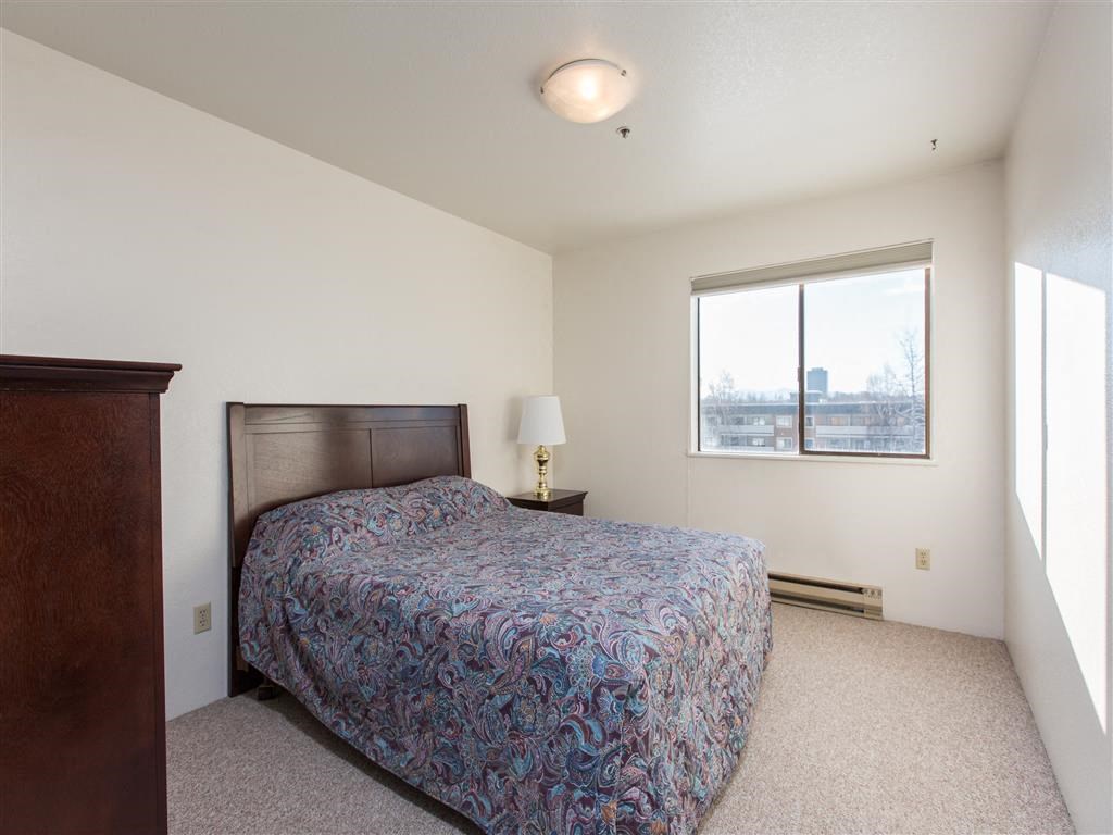 The Outlook Apartment - Bedroom Simple bedroom here at Outlook Apartments with a paisley-patterned bed, wooden headboard, and nightstand with lamp. Window shows cityscape, creating a serene atmosphere.