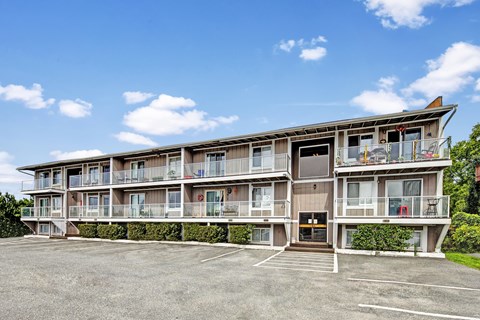 Exterior view of Arbor Pointe apartment building with balconies, large windows, and a parking lot in front under a blue sky with scattered clouds.