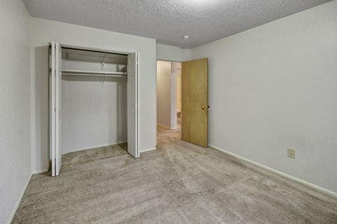 A bright bedroom at Arctic Sun Apartments with plush beige carpet and neutral walls. An open closet with double doors offers storage space. A doorway leads to the hallway and bathroom, and a ceiling light adds soft illumination, creating a comfortable and versatile room layout.