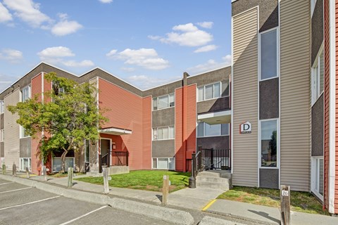 A sunny exterior view of Arctic Sun Apartments showing a two-story building with red and tan siding, large windows, and secure entryways. Small lawns and trees add greenery, and numbered parking spaces sit right outside for resident convenience. The sky is bright with scattered clouds.