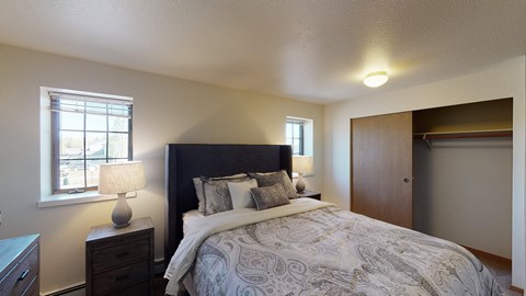 A bright Birchwood bedroom featuring a dark upholstered headboard with paisley bedding and layered pillows centered against a neutral wall. Two matching lamps on wooden nightstands sit beside the bed beneath twin windows. The room includes soft carpet and a wide closet with sliding wooden doors partially open.