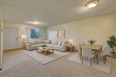 A spacious Brighton Woods living and dining area with soft neutral décor, featuring a large light gray sectional sofa, two coordinating area rugs, and a wooden coffee table. A matching dining set for six sits nearby, accented by a potted plant and simple wall art, all illuminated by warm overhead lighting and a bright window view.