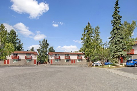 A sunny Brighton Woods community courtyard with two matching two-story apartment buildings featuring tan siding, red trim doors, and small balconies facing a wide parking area. A few cars are parked to the right, and tall evergreens and leafy trees frame the property beneath a bright blue sky dotted with soft white clouds.