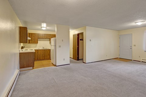 A roomy Brighton Woods living area with soft beige carpeting and cream walls flowing into an open kitchen with warm wood cabinetry and a white refrigerator. Ceiling lights brighten the space, and a front entry door and nearby hallway offer access to bedrooms and storage, creating a practical and comfortable layout.