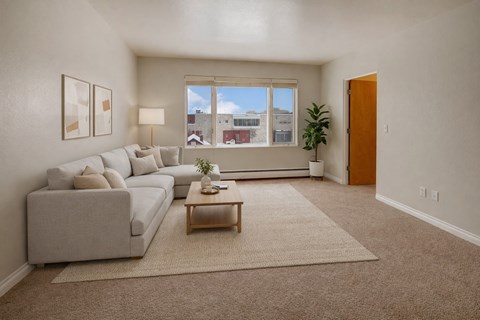A bright living room here at Century Plaza featuring a large window that fills the space with natural light and frames a city view. A soft neutral sectional sits atop a textured rug beside a wooden coffee table, paired with modern wall art, a floor lamp, and a tall potted plant creating a calm, inviting atmosphere.