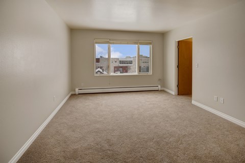 A bright living space here at Century Plaza showcasing an open layout with neutral wall tones and plush carpeting. A large picture window lets in natural light and offers a view of nearby buildings under a clear blue sky, while an adjoining doorway connects the room to the rest of the apartment.