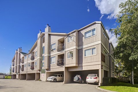 A sunny exterior view of Chugach South showing a three-story tan apartment building with small balconies, carports with parked vehicles, and trees and blue sky in the background.