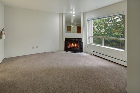 A cozy living room here at Chugach South featuring light walls, beige carpeting, a lit wood-burning fireplace with a mirror above it, a large window overlooking green trees, and a fire extinguisher mounted on the wall.