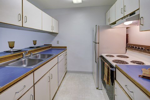 A bright kitchen here at Chugach South featuring clean white cabinetry with wood trim, vivid blue countertops, and matching upper and lower storage on both sides of the room. Stainless steel double sinks, a classic electric stove, and a full-size refrigerator sit beneath soft wall lighting, creating a tidy and welcoming cooking space.