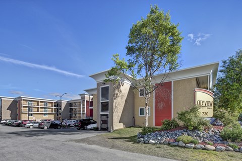A sunny exterior view of City View Apartment Homes showcasing modern multi-story buildings with bold red and tan accents, balconies, and well-kept landscaping framed by rocks, shrubs, and trees. A clear blue sky, parked cars, and the welcoming entrance sign create a bright and inviting first impression of the community.