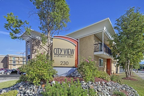 A welcoming front view of City View Apartment Homes featuring the large entrance sign surrounded by colorful landscaping with rocks, shrubs, and blooming flowers. Modern tan buildings with balconies rise behind it, and a bright blue sky adds a cheerful backdrop to this well-kept community.