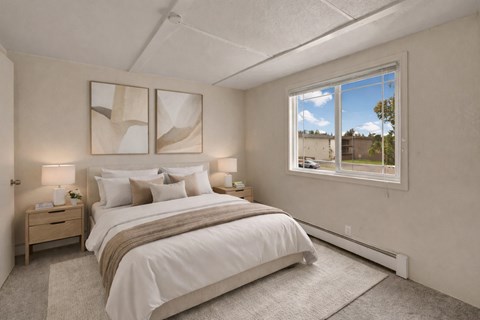 A bright, inviting bedroom here at College View featuring soft neutral tones, plush carpet flooring, and a large window that brings in natural light. The space includes a comfortable bed with layered white bedding, matching nightstands with lamps, and modern wall art, creating a calm, functional retreat with a clean, welcoming layout.