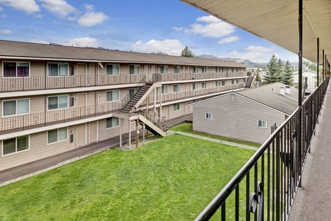 A bright courtyard view here at College View showcasing multi-story apartment buildings with exterior walkways, railings, and staircases overlooking a well-maintained green lawn. Neutral siding, large windows, and open corridors create a clean, welcoming community layout with mountain views and blue skies visible in the background.