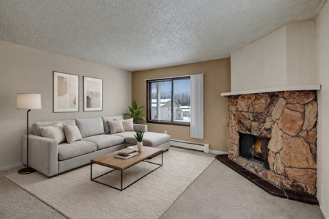 A cozy living room here at Conifer Grove featuring a light gray sectional sofa with neutral pillows, a simple wooden coffee table, and a soft area rug that defines the seating space. A large window brings in natural light, while a striking stone fireplace adds warmth and character to the room.