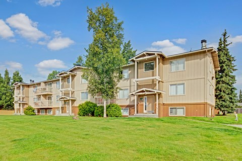 A sunny exterior view here at Conifer Grove showcasing a multi-story apartment building with neutral siding, private balconies, and large windows, surrounded by well-maintained green lawns, mature trees, and a bright blue sky that highlights the community’s peaceful residential setting.