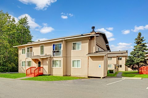 A bright exterior view here at Conifer Grove featuring a well-kept apartment building with neutral-toned siding, white-trimmed windows, and small private balconies, complemented by red entry railings, paved walkways, nearby parking, and surrounding greenery under a clear blue sky.