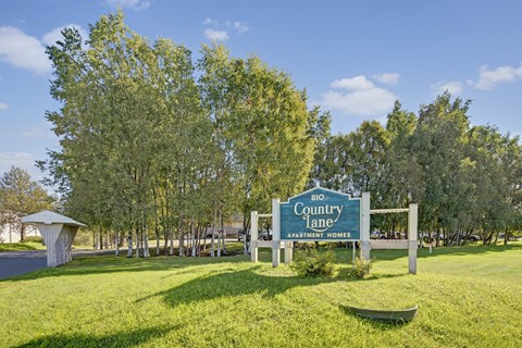 A welcoming community sign here at Country Lane displaying the Country Lane Apartment Homes name and address, set against a landscaped green lawn with mature trees and a clear blue sky, marking the entrance to the apartment community.