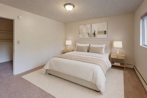 A bright bedroom here at Country Lane featuring a queen-sized bed with white bedding and neutral accent pillows, matching nightstands with lamps, soft carpeting, an area rug under the bed, abstract wall art, and an open closet providing simple storage and a calm, comfortable layout.