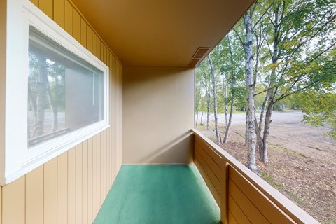 A covered balcony here at Country Lane offering a private outdoor space with neutral siding, sturdy railing, and a soft green floor surface, providing a peaceful spot to enjoy fresh air and views of surrounding trees while extending the apartment’s living space.