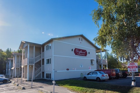 A welcoming exterior view of Driftwood featuring light-colored apartment buildings with covered stairways and balconies, surrounded by mature trees, open green space, and on-site parking, creating a quiet residential setting with a well-maintained community atmosphere.