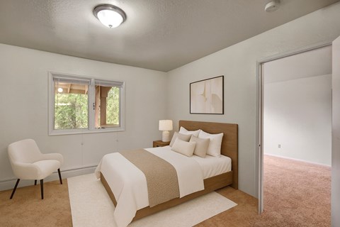A calm bedroom here at Driftwood featuring soft carpet flooring, a spacious layout, a cozy bed with neutral bedding and accent pillows, a warm wood headboard, bedside table with lamp, natural light from the window, and leading into an adjacent room, creating a comfortable and inviting retreat.