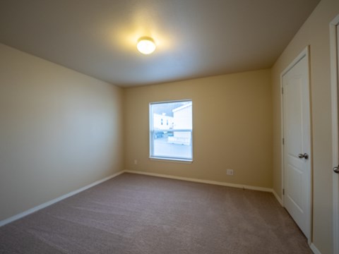 A bright, carpeted bedroom here at Four Seasons featuring soft neutral walls, a large window that brings in natural light, and a simple overhead ceiling fixture. The open layout offers flexible space for bedroom furniture, with a clean finish, white trim, and a nearby closet door creating a comfortable and inviting retreat.