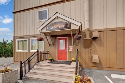 A welcoming leasing office entrance here at The Greenbriar featuring a red front door beneath a covered entryway, decorative hanging flower baskets, and a clearly marked leasing office sign. The image highlights clean steps with railings, potted plants for added curb appeal, and neutral-toned exterior siding that creates a friendly, approachable first impression.
